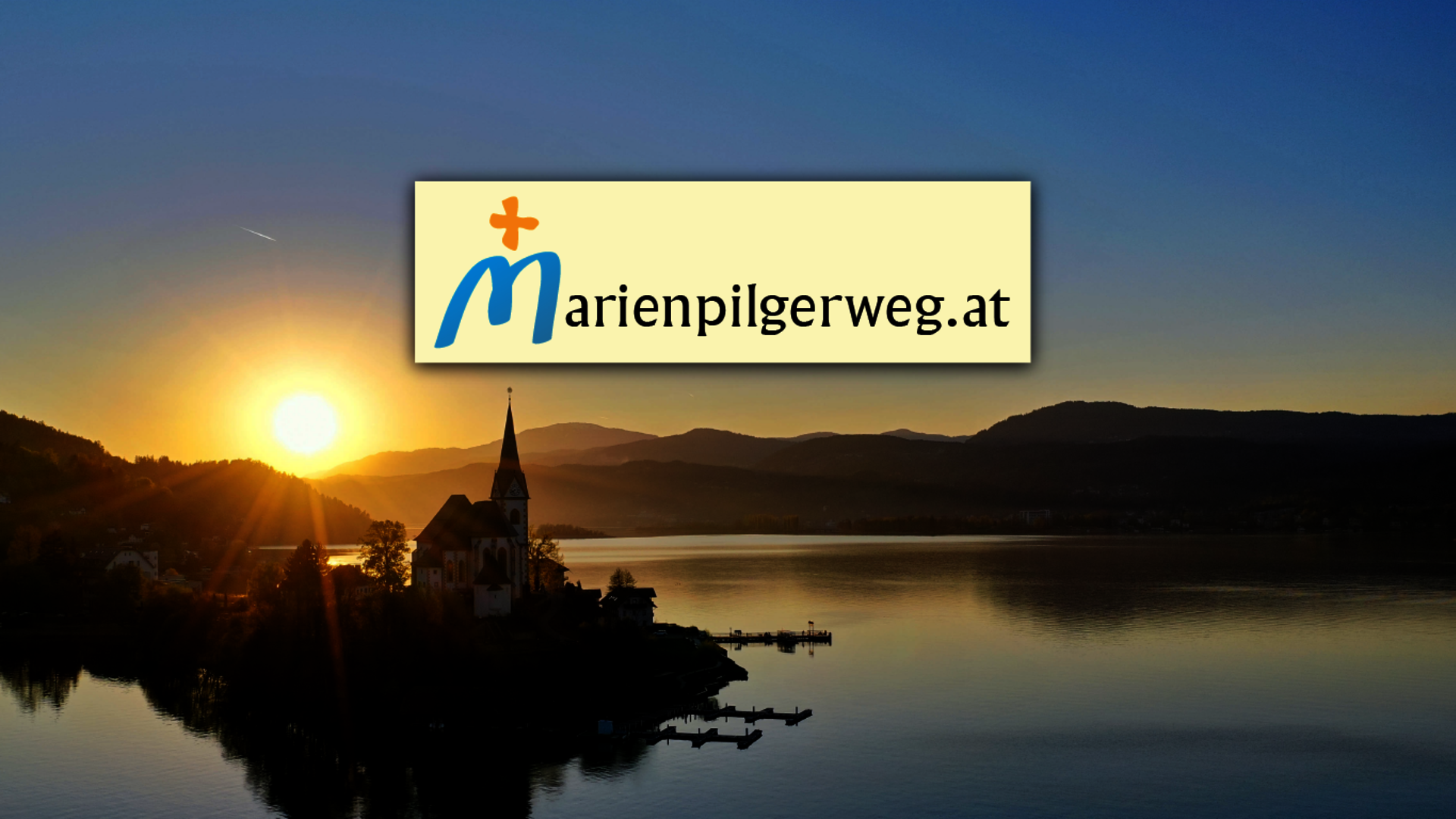 <p>Aufnahme der Kirche Maria Wörth im Sonnenuntergang mit Logo des Marienpilgerweges.</p>